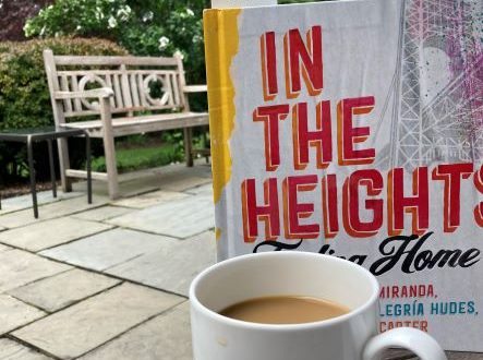 image of in the heights finding home with a full cop of coffee in front of a treelined backgroud with an umbrella and a bench.